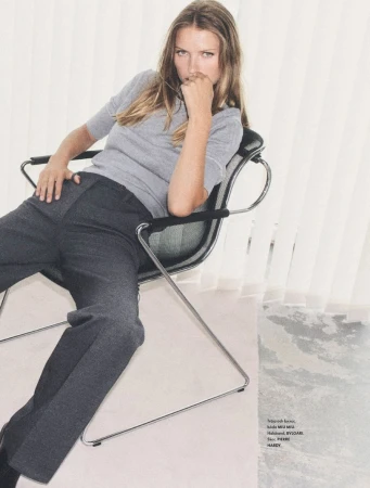 A young adult woman is seated on a chair in a studio, wearing a gray sweater and gray slacks, with a window visible in the background.