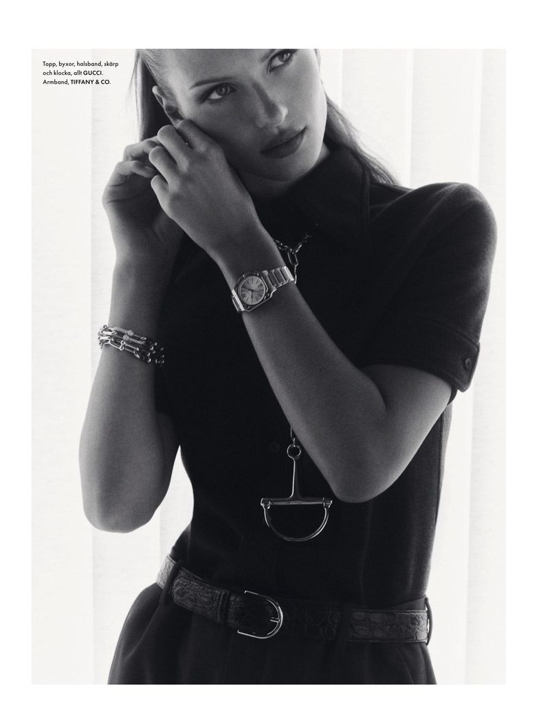 Monochrome fashion shot of a woman adjusting an earring, wearing a dress with a belt, and jewelry, against a bright, neutral backdrop.