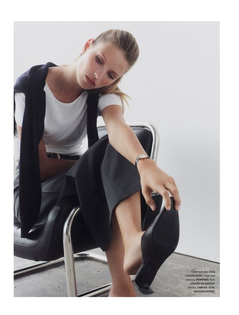 Fashion shot of a blond woman wearing a white shirt and dark pants, seated on a metal chair while touching a black shoe.