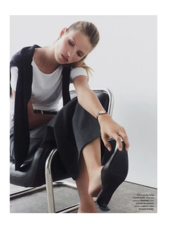 Fashion shot of a blond woman wearing a white shirt and dark pants, seated on a metal chair while touching a black shoe.