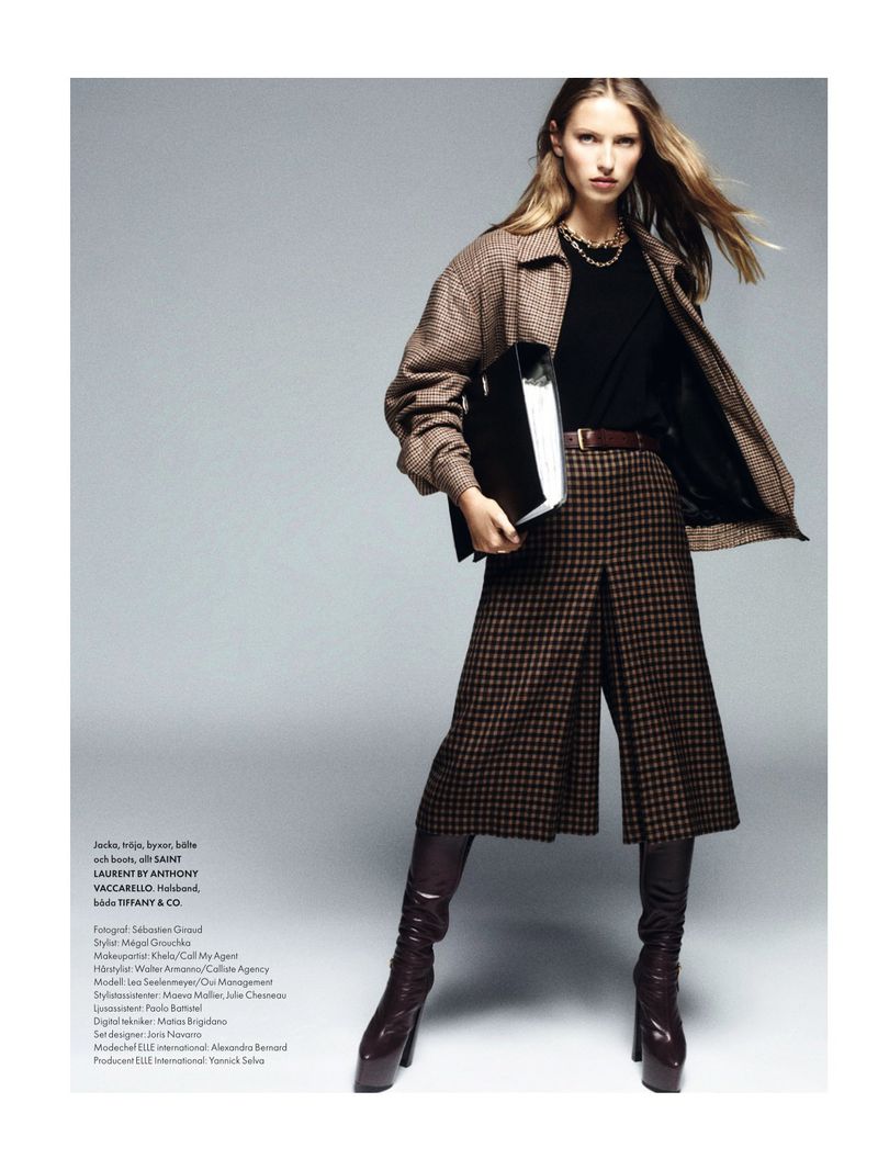 Fashion editorial shot of a blonde woman in patterned culottes, a brown jacket, and knee-high boots, posing with a black folder.
