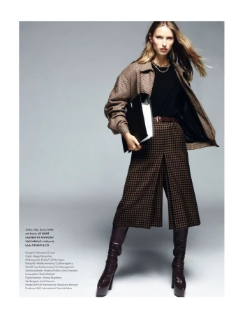 Fashion editorial shot of a blonde woman in patterned culottes, a brown jacket, and knee-high boots, posing with a black folder.