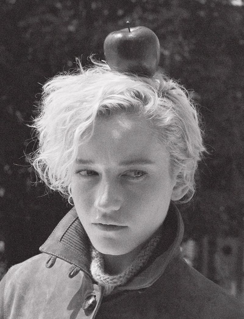 Black and white portrait of a woman with an apple on her head, wearing a jacket and sweater, set against an outdoor backdrop.
