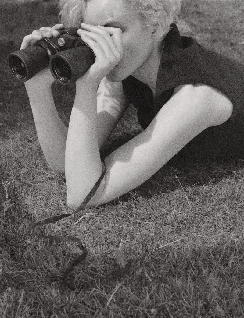Adult female looking through binoculars outdoors in black and white. Woman with short blonde hair lies prone in the grass.