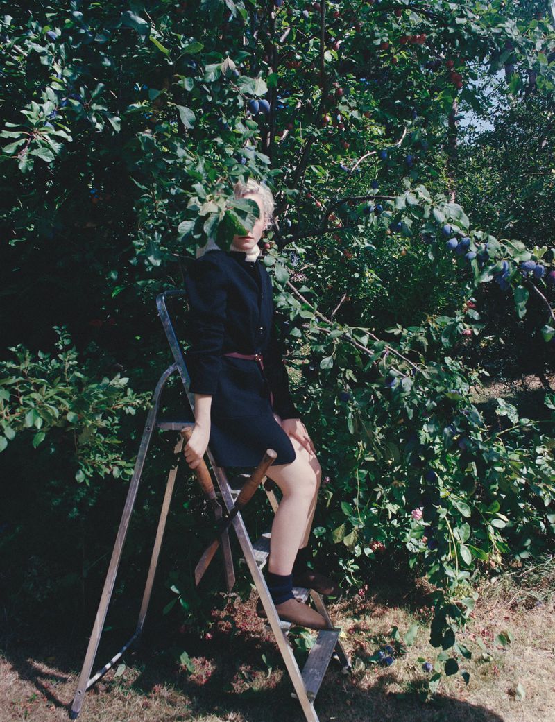 Woman in a dark dress sitting on a metal ladder outdoors among leafy plum trees with visible fruit on a sunny day, holding leaves.