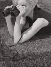 Blonde woman lying in grass looking through black binoculars for Pop Magazine editorial
