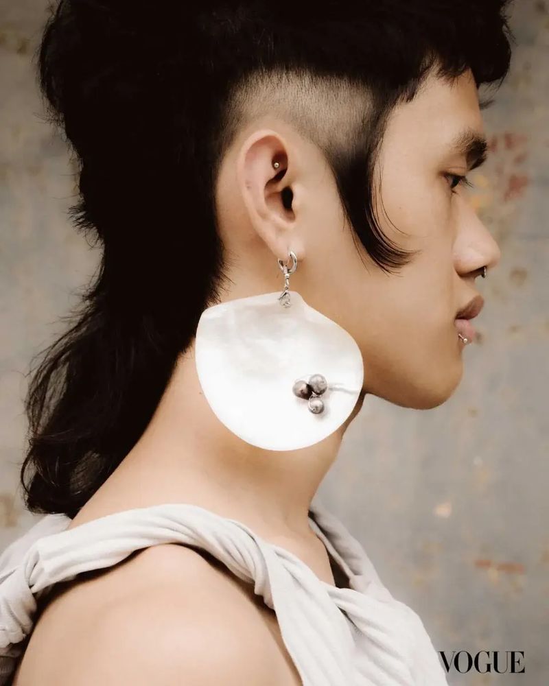 Close-up side-view portrait of an adult with a mullet, wearing a large seashell earring, posing against a textured grey background.