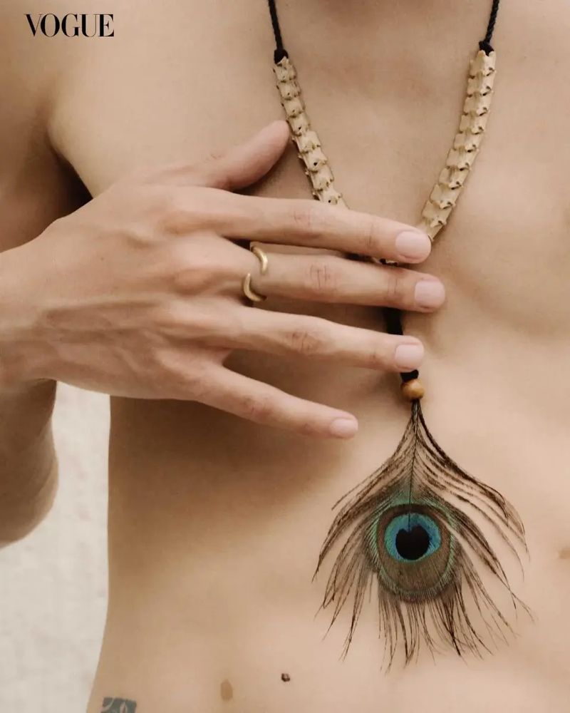 Close-up fashion shot of a shirtless person with a bone and peacock feather necklace, gold ring, against a neutral background, Vogue logo.