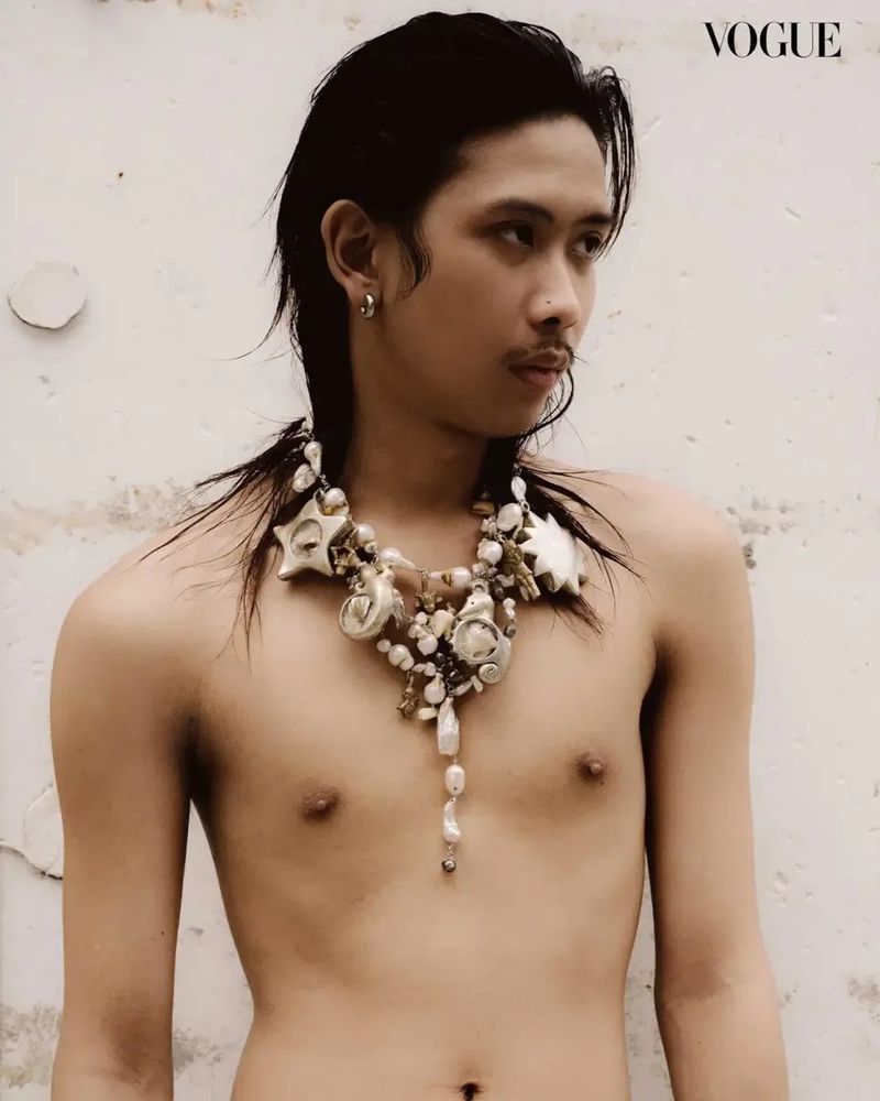 Shirtless adult male poses against a white wall wearing a shell necklace in a medium shot with editorial Vogue logo.