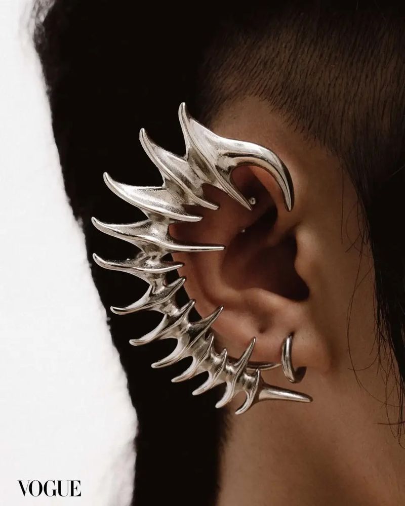Close-up of an adult with short dark hair, wearing a dramatic silver ear cuff earring in a studio with grey and white background.