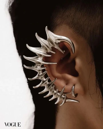 Close-up of an adult with short dark hair, wearing a dramatic silver ear cuff earring in a studio with grey and white background.