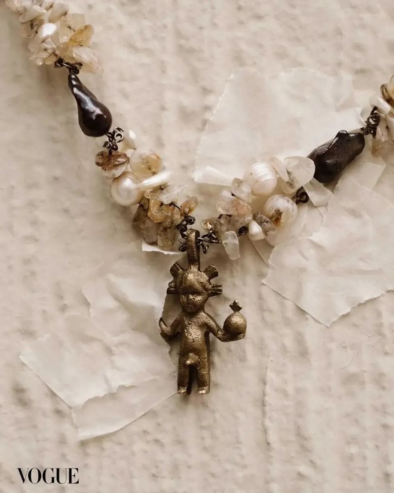 Close-up of a sculptural bronze pendant of a human figure holding a sphere, hanging on a necklace of baroque pearls and stones.