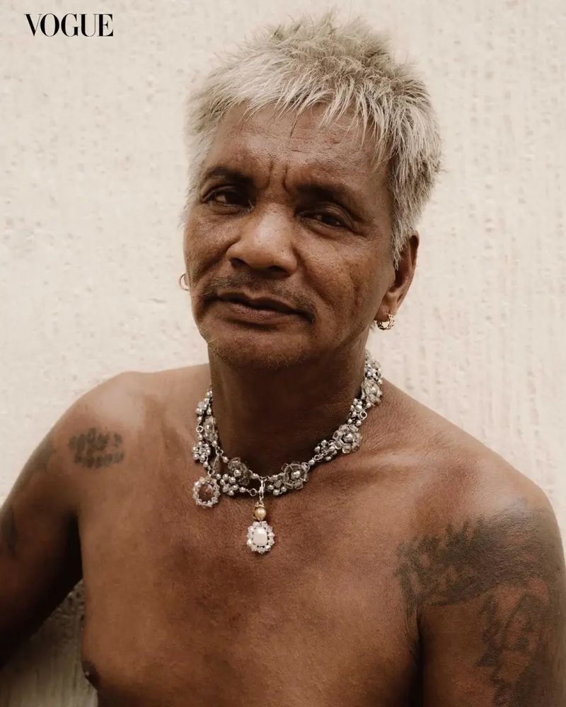 Close-up portrait of a mature adult man with gray hair, wearing a decorative silver necklace, set against a neutral background.