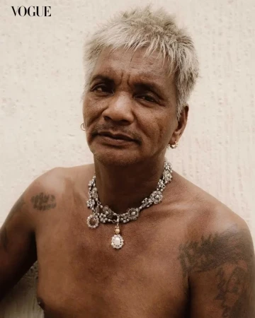 Close-up portrait of a mature adult man with gray hair, wearing a decorative silver necklace, set against a neutral background.