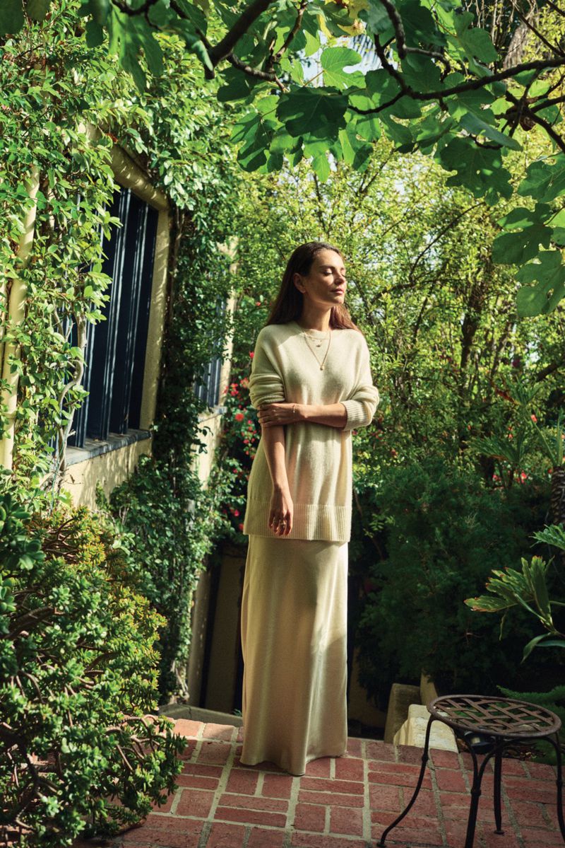 Woman stands in sunlight wearing a matching beige sweater and skirt in an outdoor setting filled with lush green foliage.