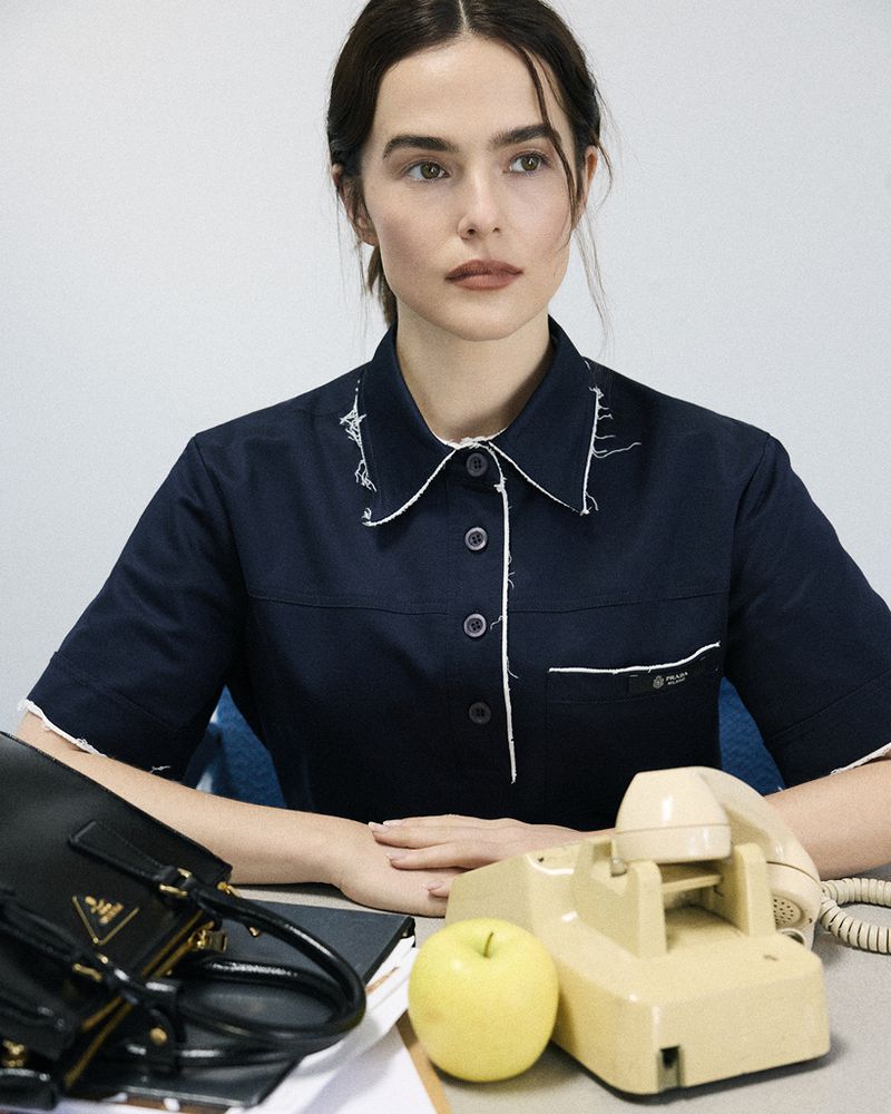 Adult female in a navy shirt sitting at a desk with a landline phone, apple, and Prada handbag, with a white and light blue background.