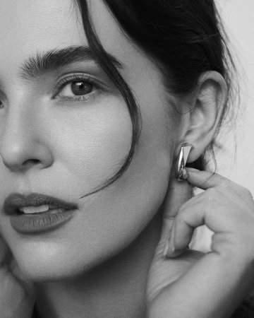 Close-up black and white portrait of actor Zoey Deutch wearing and touching a sculptural metal earring against a studio backdrop.