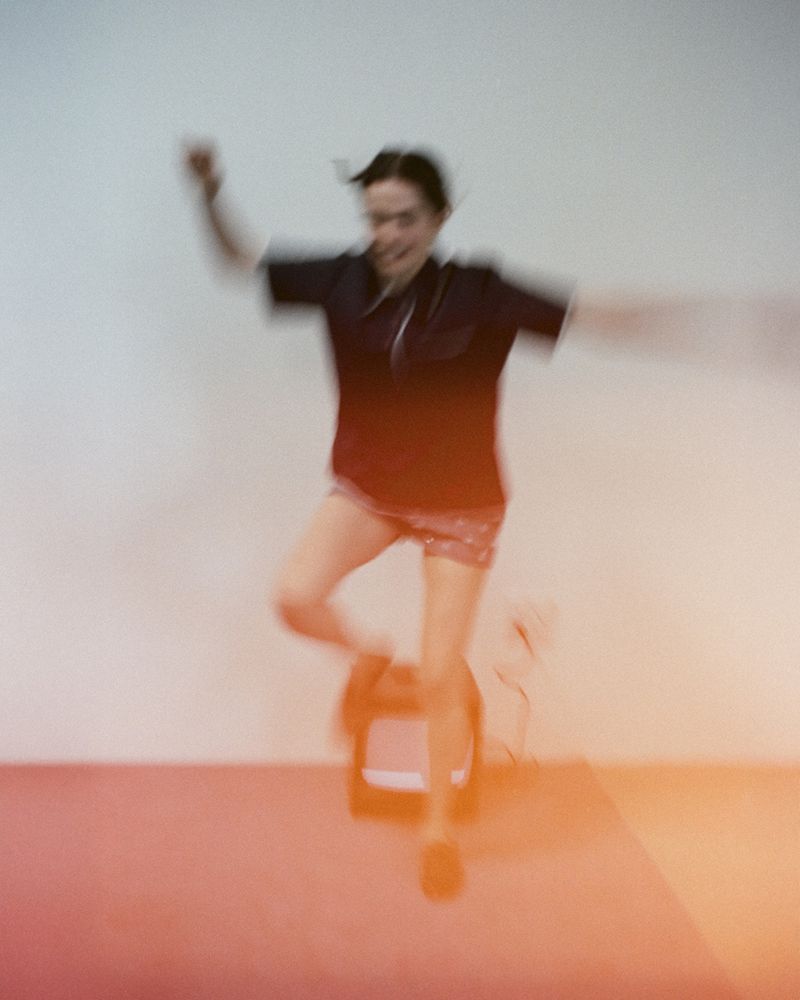 Blurred action shot of a woman in shorts and short-sleeve shirt, jumping in an indoor setting with a red floor against a white wall.
