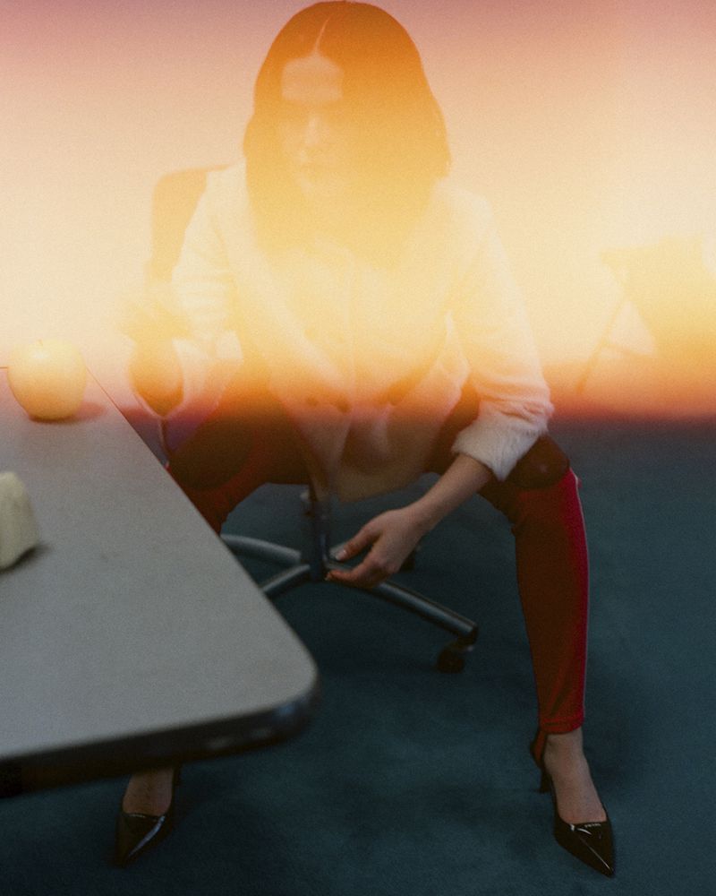 Female fashion model sits at a desk in a brightly lit room, wearing a white top, red pants, and black heels. 
