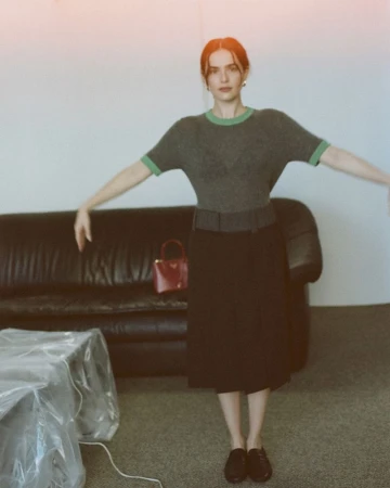 Zoey Deutch standing with outstretched arms wearing a grey knit top and black midi skirt in front of a dark leather couch.