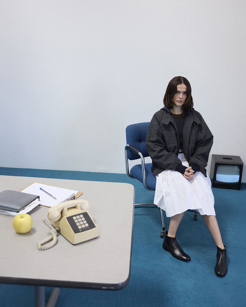 A young woman sits in a blue office chair with an old television in the background and a tan rotary phone on a desk.