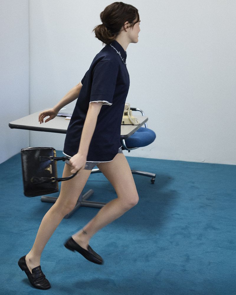 Woman walking quickly across a blue carpeted room carrying a black handbag, wearing a dark polo shirt and loafers.