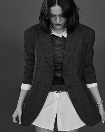A stylish woman poses in a dark-grey plaid blazer and white collared shirt, looking down in a black-and-white studio fashion shot.