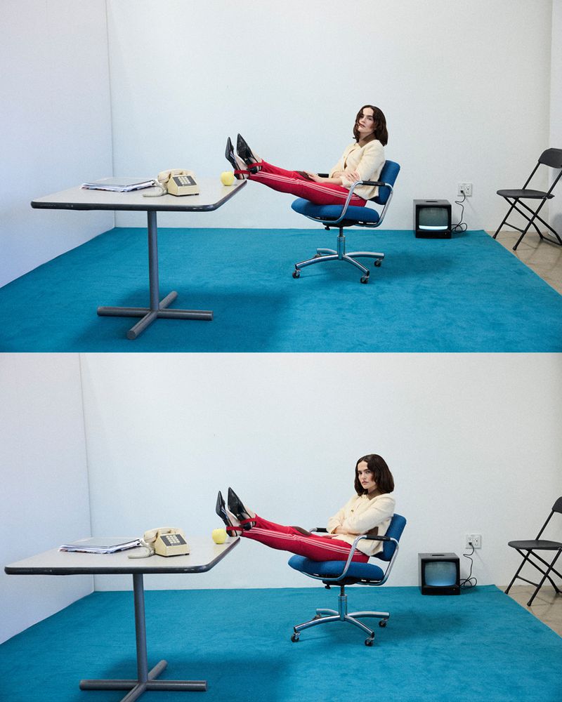 A woman in red pants sits in a blue office chair with her feet up on a desk in a blue and white office, with a retro television set.