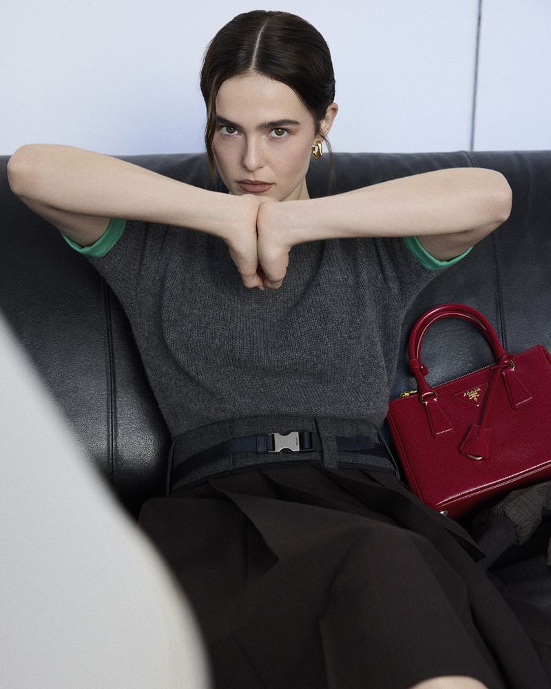 Portrait of a stylish adult woman with dark hair and a gray top posed on a black couch with a red handbag next to her.