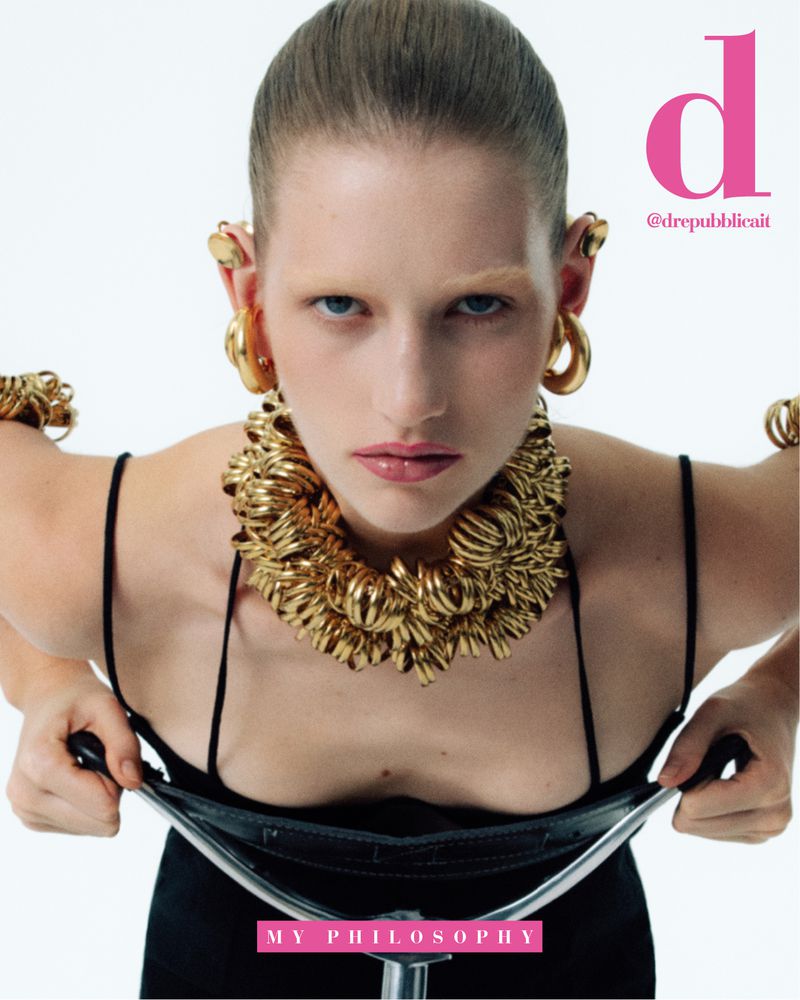 Blonde woman with statement gold jewelry posing with vintage bicycle, wearing black dress against white background in fashion editorial.