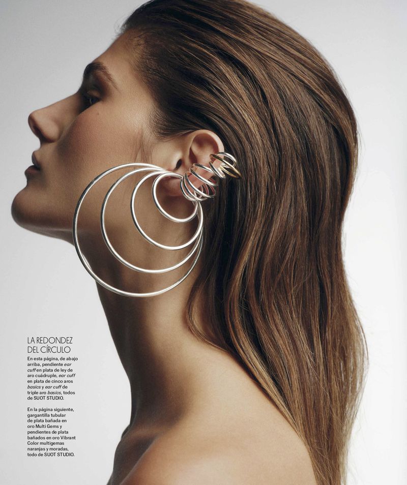 Profile of a woman with brown hair wearing large silver hoop earrings and a gold ear cuff against a plain light background.