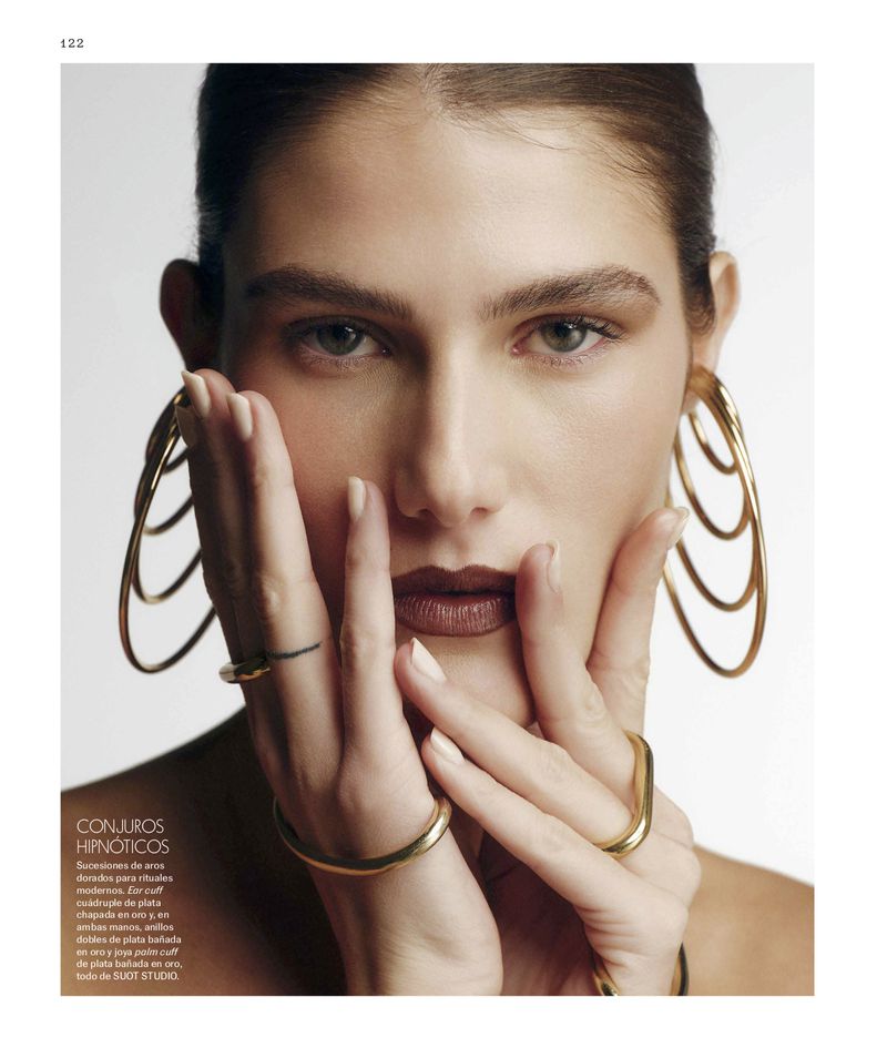 Close-up of a woman with brown hair and brown lipstick, wearing gold jewelry, and posing with her hands up near her face.