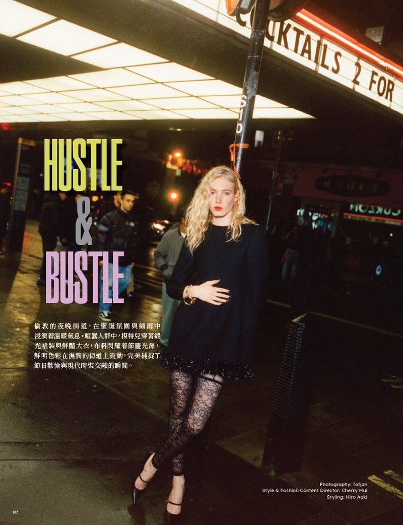 A fashion shot shows a woman posing in a black mini dress and lace tights on a city street at night under a sign for cocktails.