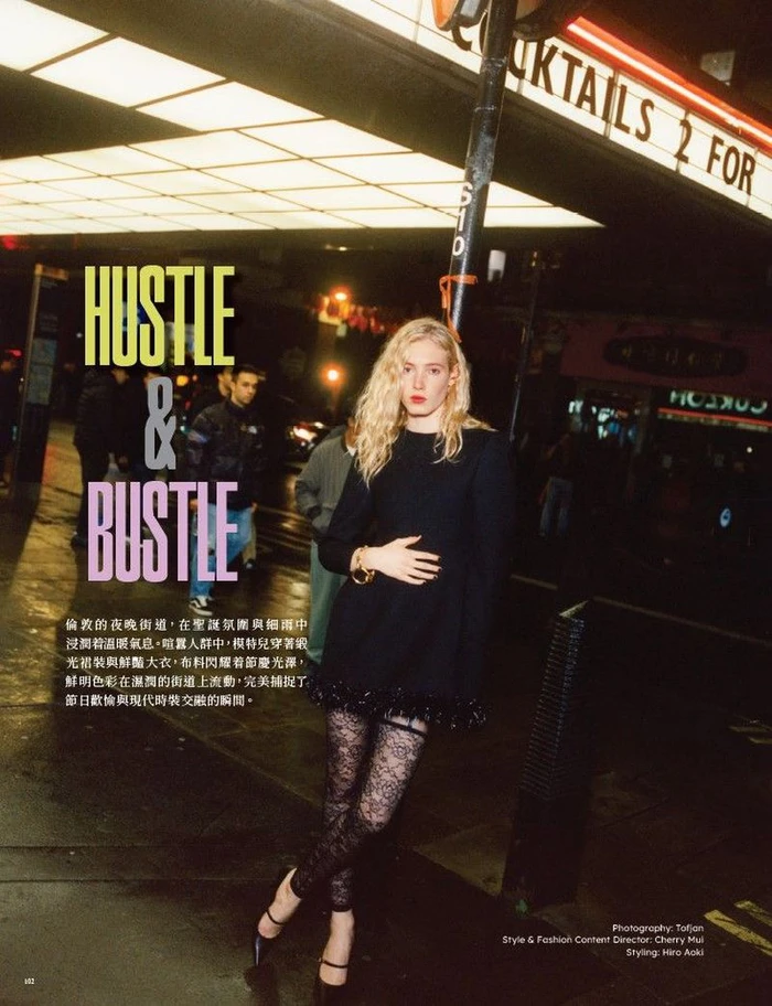 Blonde woman wearing a black dress and patterned tights posing on a wet city street at night under neon signs
