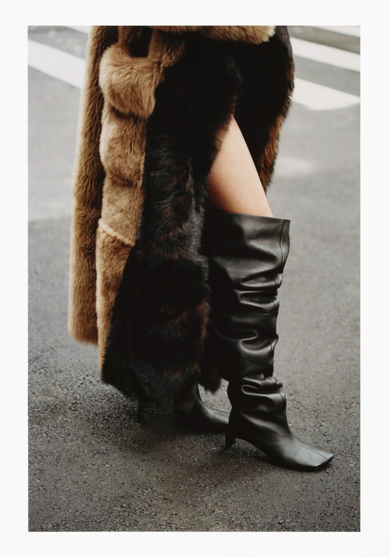 Fashion forward photograph with a woman wearing a long coat with fur trim and a pair of black leather boots walking on a street.
