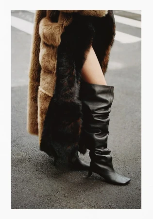 Fashion forward photograph with a woman wearing a long coat with fur trim and a pair of black leather boots walking on a street.