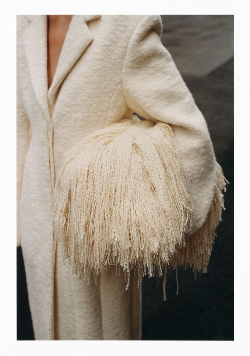 Close-up of a person wearing a cream-colored coat with fringed sleeves in front of a dark background, minimalist fashion concept.