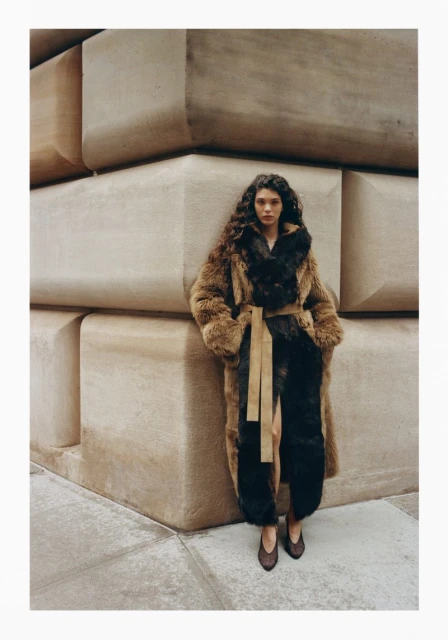 A fashionable adult woman with curly hair poses outside wearing a fur coat and leather belt, standing next to large concrete blocks.