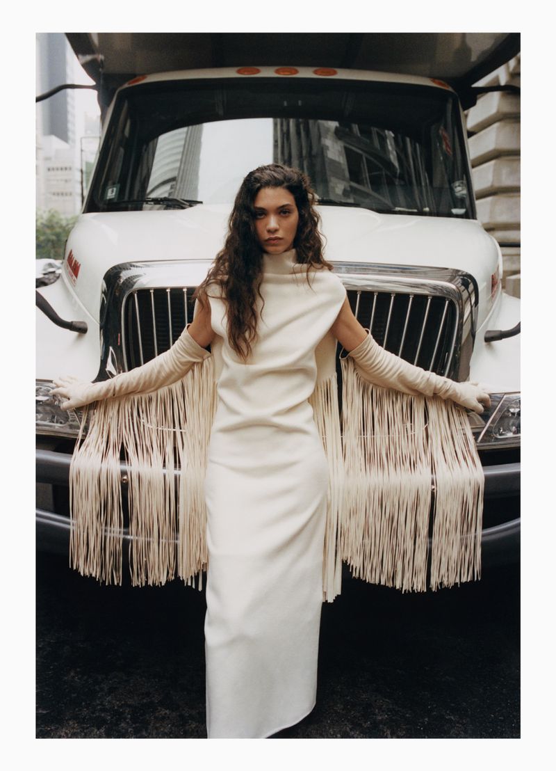Woman in long fringe dress and matching gloves posing against the front of a large white truck in an urban setting.