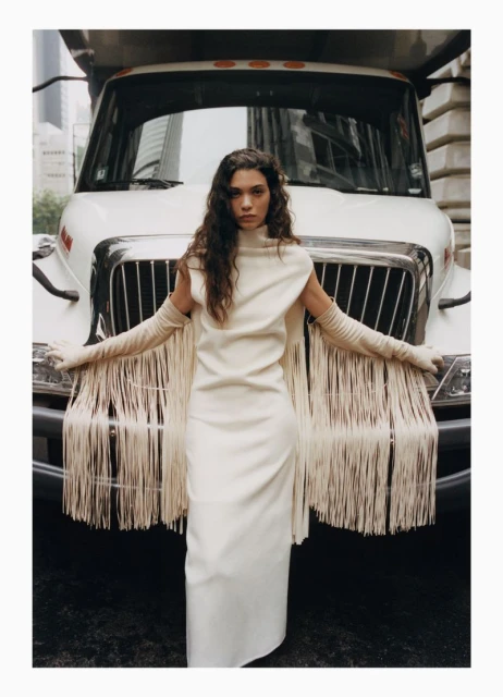 Woman in long fringe dress and matching gloves posing against the front of a large white truck in an urban setting.