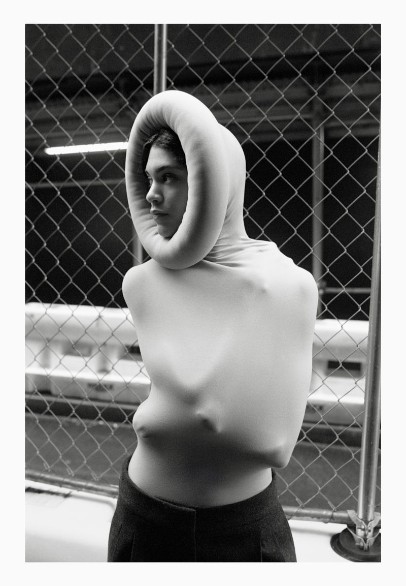 A woman in a close-up studio shot wearing a sculptural white garment and dark pants poses in front of a metal fence for a fashion editorial.