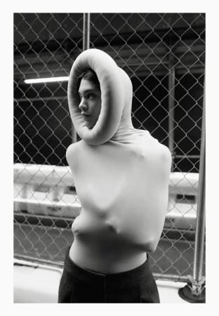 A woman in a close-up studio shot wearing a sculptural white garment and dark pants poses in front of a metal fence for a fashion editorial.