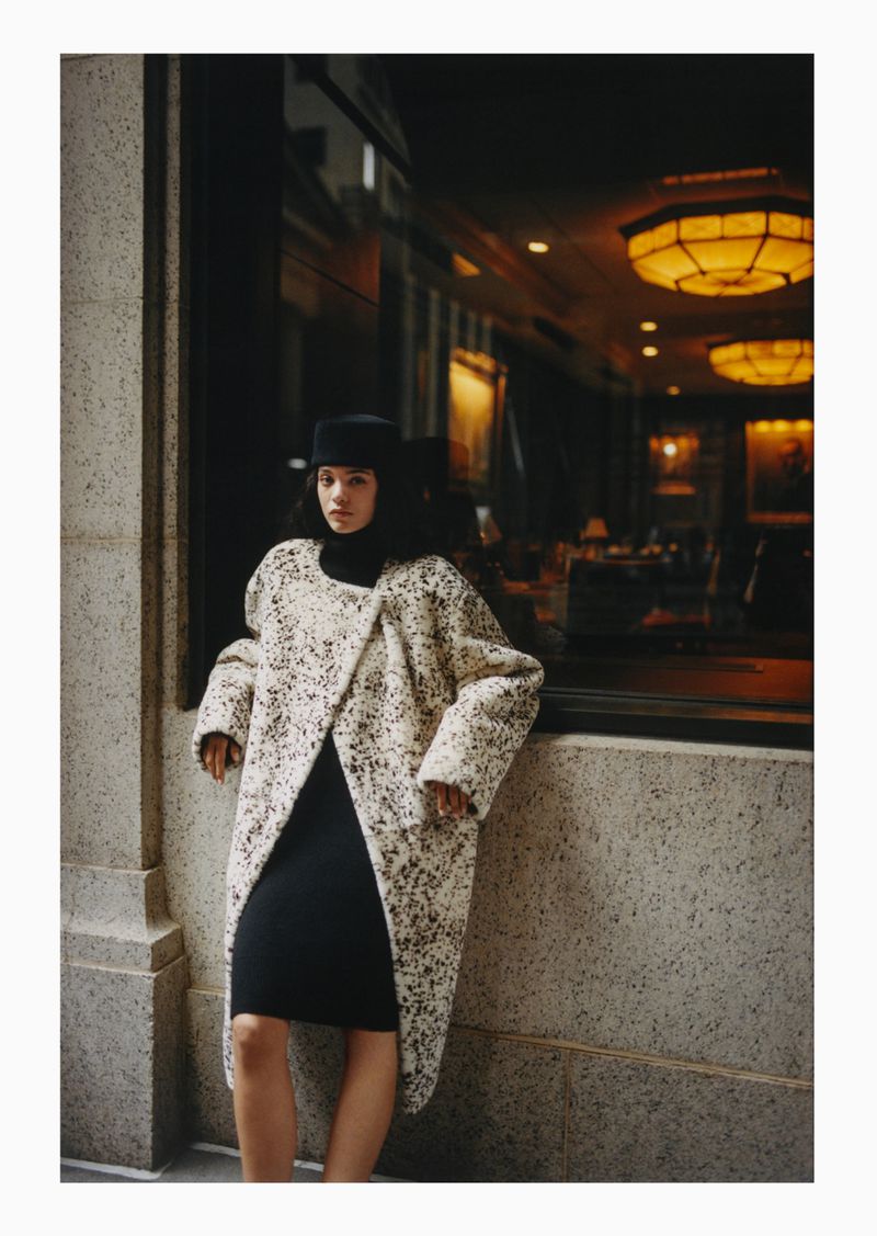 A woman in a speckled coat and black dress leans against a wall next to a window, showcasing a blend of modern fashion and urban ambiance.