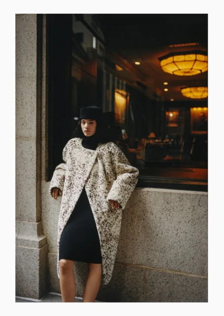 A woman in a speckled coat and black dress leans against a wall next to a window, showcasing a blend of modern fashion and urban ambiance.