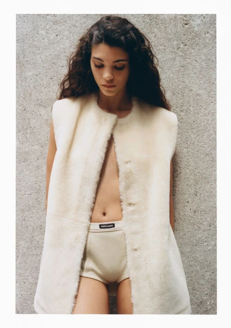 A woman poses in a photo studio wearing underwear and a sleeveless fur coat. Fashion editorial shot of a slender female model.