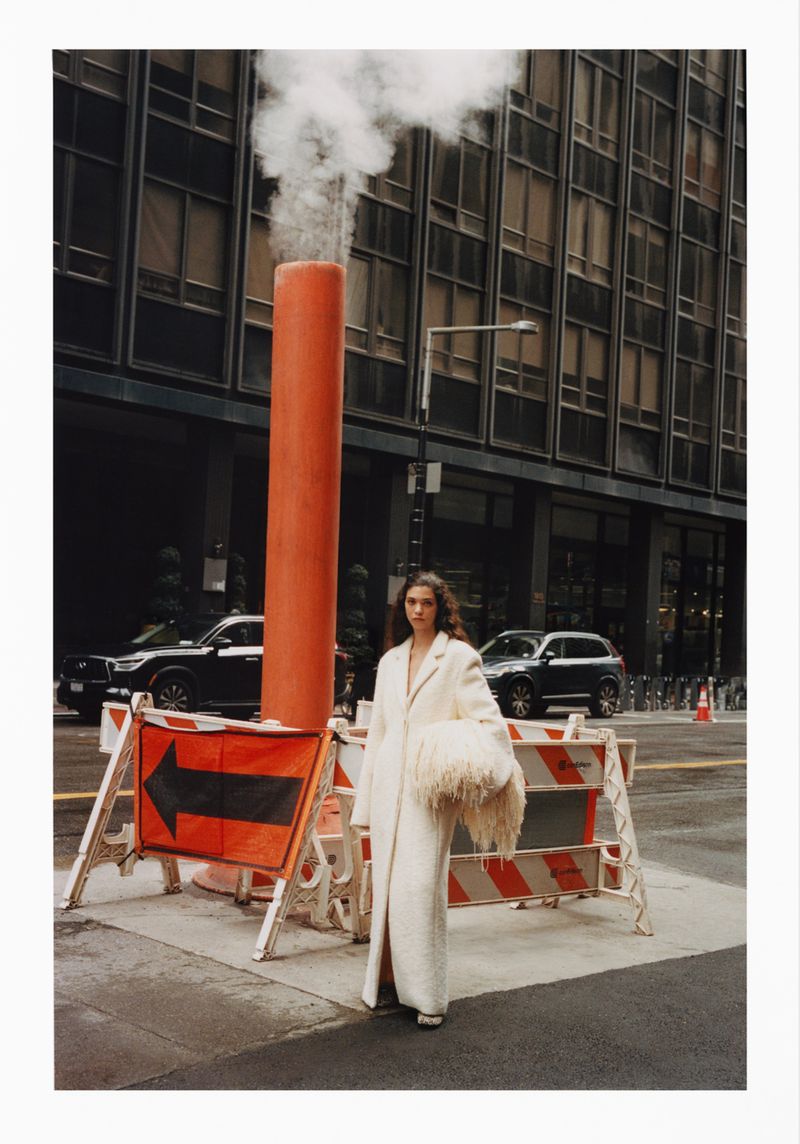Adult female fashion model poses in a textured white coat, holding a fuzzy bag on a city street with roadwork barriers and skyscrapers.