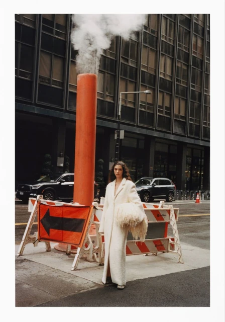 Adult female fashion model poses in a textured white coat, holding a fuzzy bag on a city street with roadwork barriers and skyscrapers.