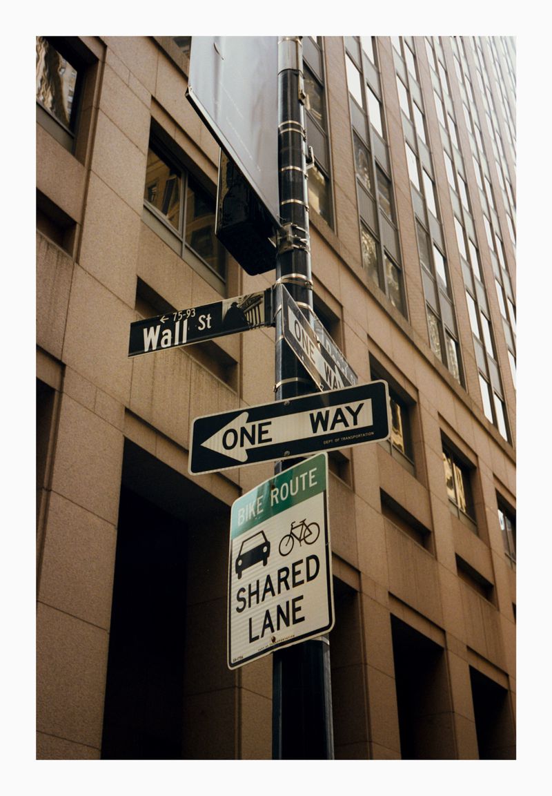 Street signs indicating Wall Street and a bike route in an urban setting, with a tall building in the background on a sunny day.
