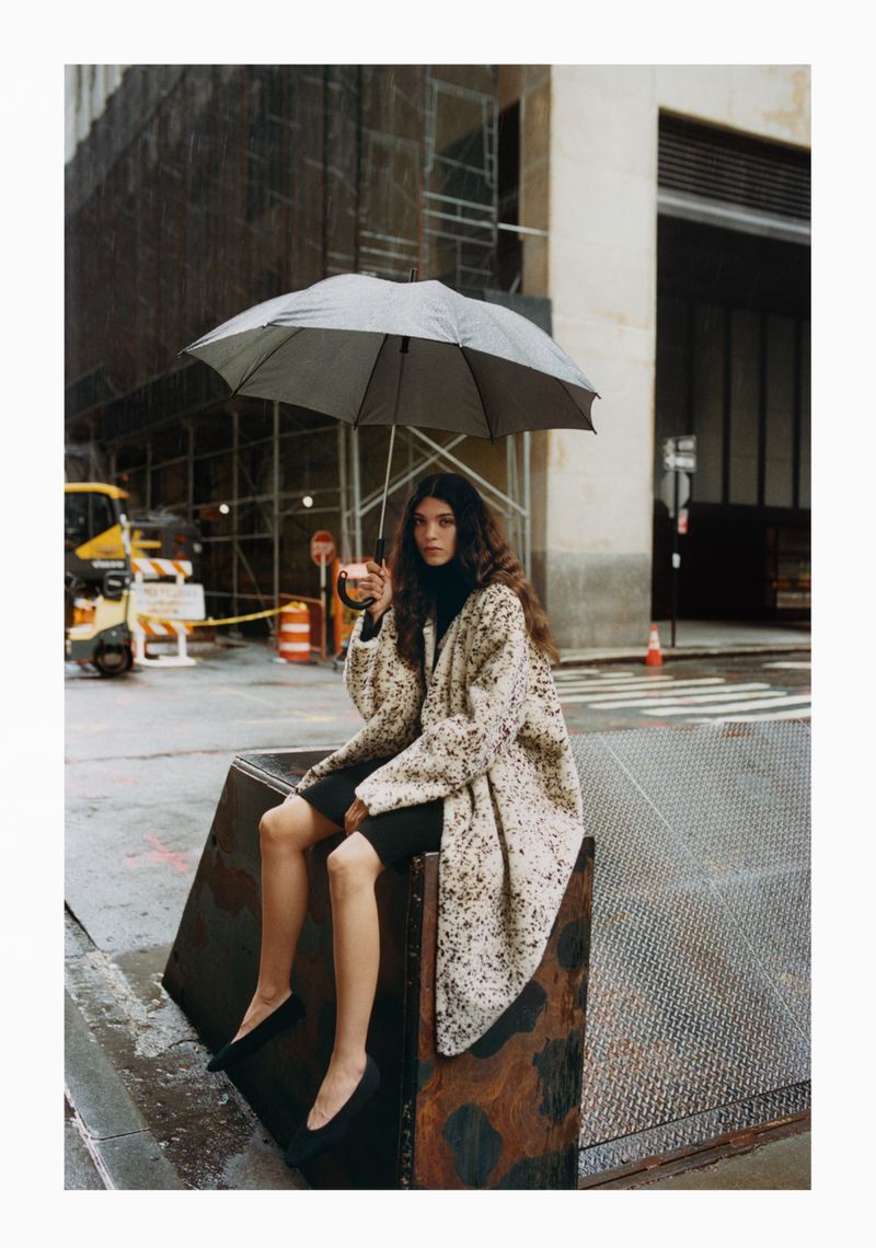 A woman with curly hair sits in a city street scene on a metal structure holding an umbrella while wearing a speckled coat.