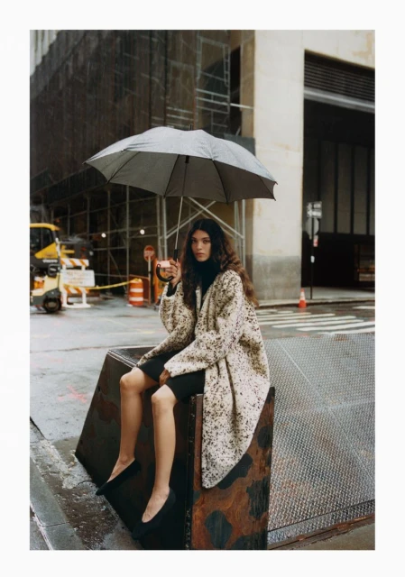A woman with curly hair sits in a city street scene on a metal structure holding an umbrella while wearing a speckled coat.